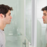 side view of a tensed young man looking at self in mirror in the bathroom
